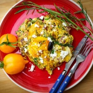 Feta and Spinach Frittata served on a red plate with two yellow tomatoes on the side, plus a blue fork and knife set