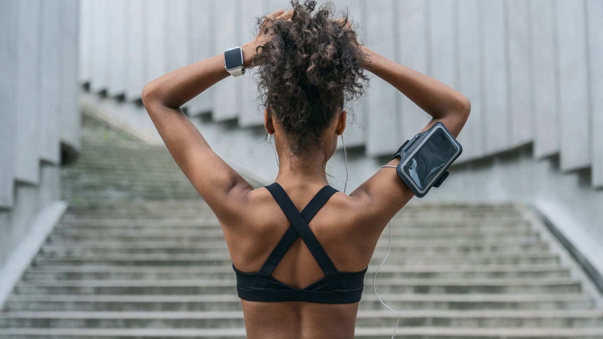 Woman in athletic clothing typing up hair and facing outdoor stairs