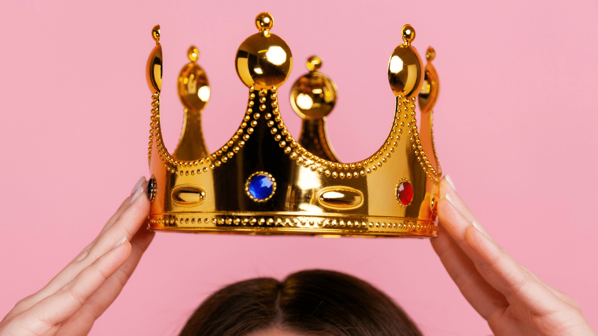 Gold crown held over head on pink background