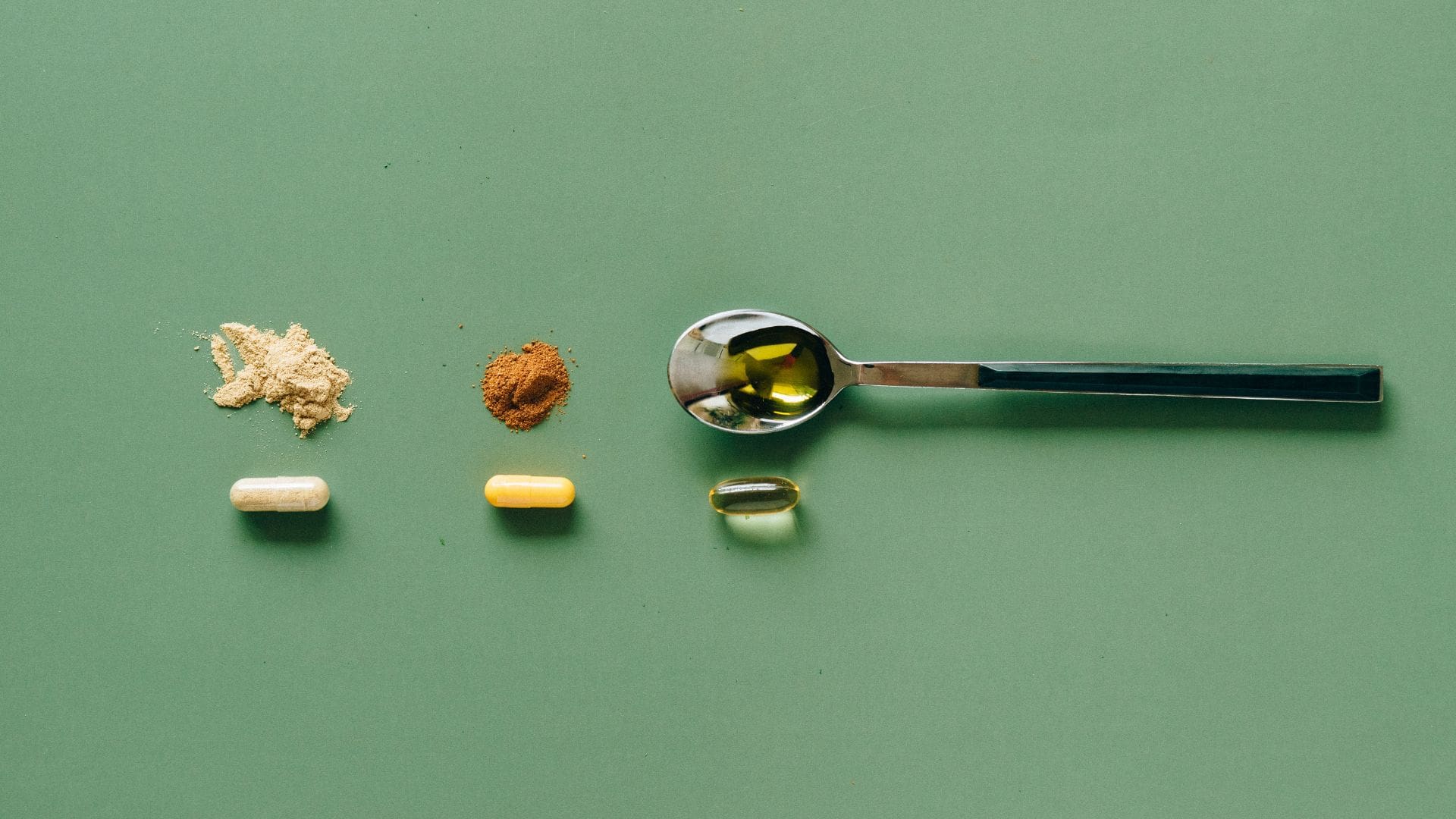 Three capsules next to powders with a spoon against green backdrop