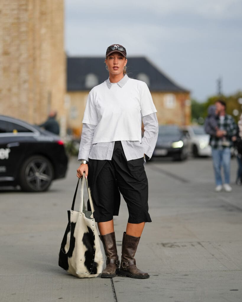 COPENHAGEN, DENMARK - AUGUST 06: A guest wears a white short-sleeved top layered over a black and white striped button-up shirt with visible cuffs and collar. Black knee-length shorts are paired with brown leather mid-calf boots featuring buckles. Accessories include a black and white cowhide / cow print pattern fur tote bag, a black cap from New Era. Sunglasses rest on the cap, and a silver watch is visible on the wrist, outside Gestuz, during Copenhagen Fashion Week, on August 06, 2025 in Copenhagen, Denmark