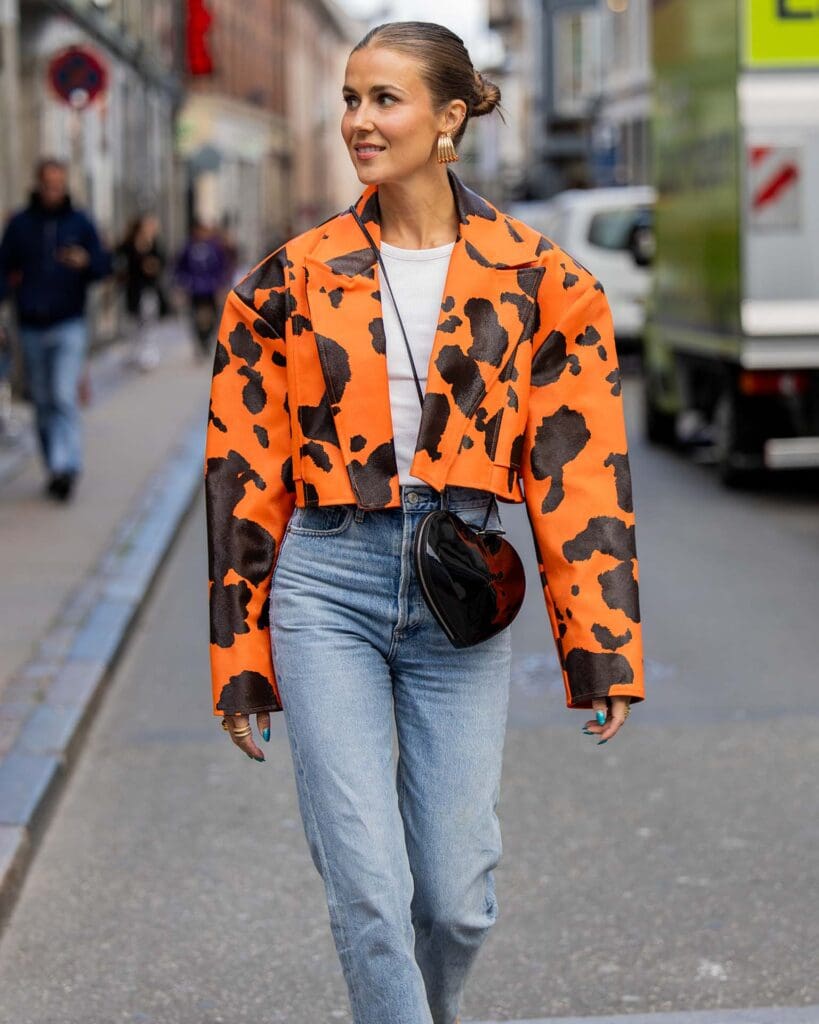 COPENHAGEN, DENMARK - AUGUST 08: Nina Sandbech wears denim jeans, black heat shaped bag, orange black bag with cow print, yellow heels, golden earrings during the Copenhagen Fashion Week Spring/Summer 2024 on August 08, 2023 in Copenhagen, Denmark.