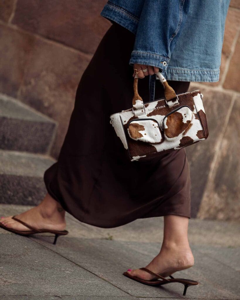 COPENHAGEN, DENMARK - AUGUST 08: Elise Marie Louise wears long brown maxi dress, blue denim jacket from Loewe, brown and white cow print hand bag and brown high heels outside the Rotate show during day four of the Copenhagen Fashion Week (CPHFW) SS25 on August 08, 2024 in Copenhagen, Denmark.